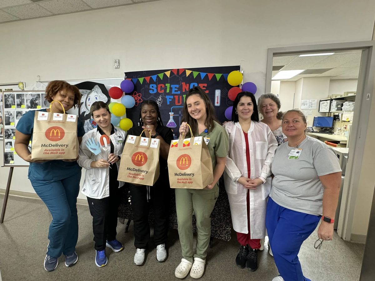 Group of six women standing together in an office setting, smiling, holding McDonald’s takeout bags labeled McDelivery.
