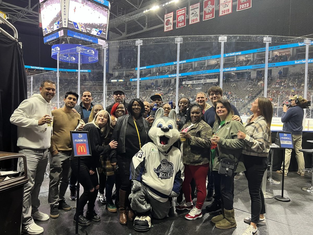 Group of people posing for a photo inside a hockey arena, with a person in a cartoon lion mascot costume.