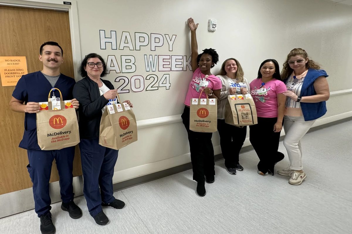 Six people standing in a hallway holding McDonald’s takeout bags in front of a sign that says 'HAPPY CLAB WEEK'.
