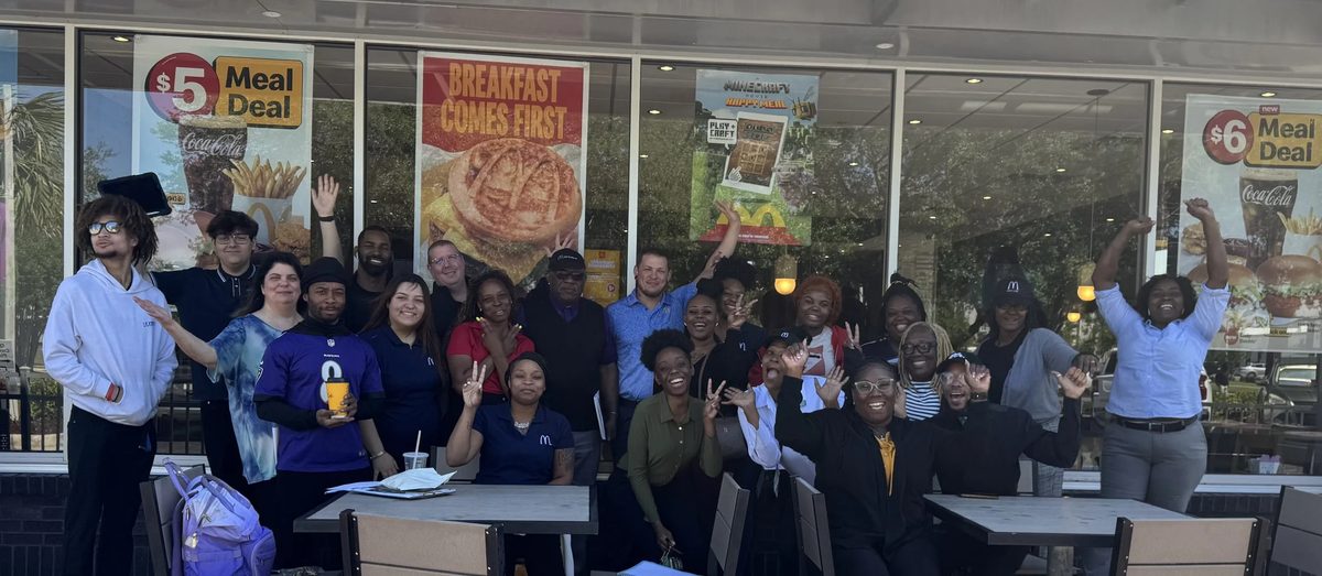 A group of people standing and sitting outside a McDonald’s restaurant, smiling and making various gestures.