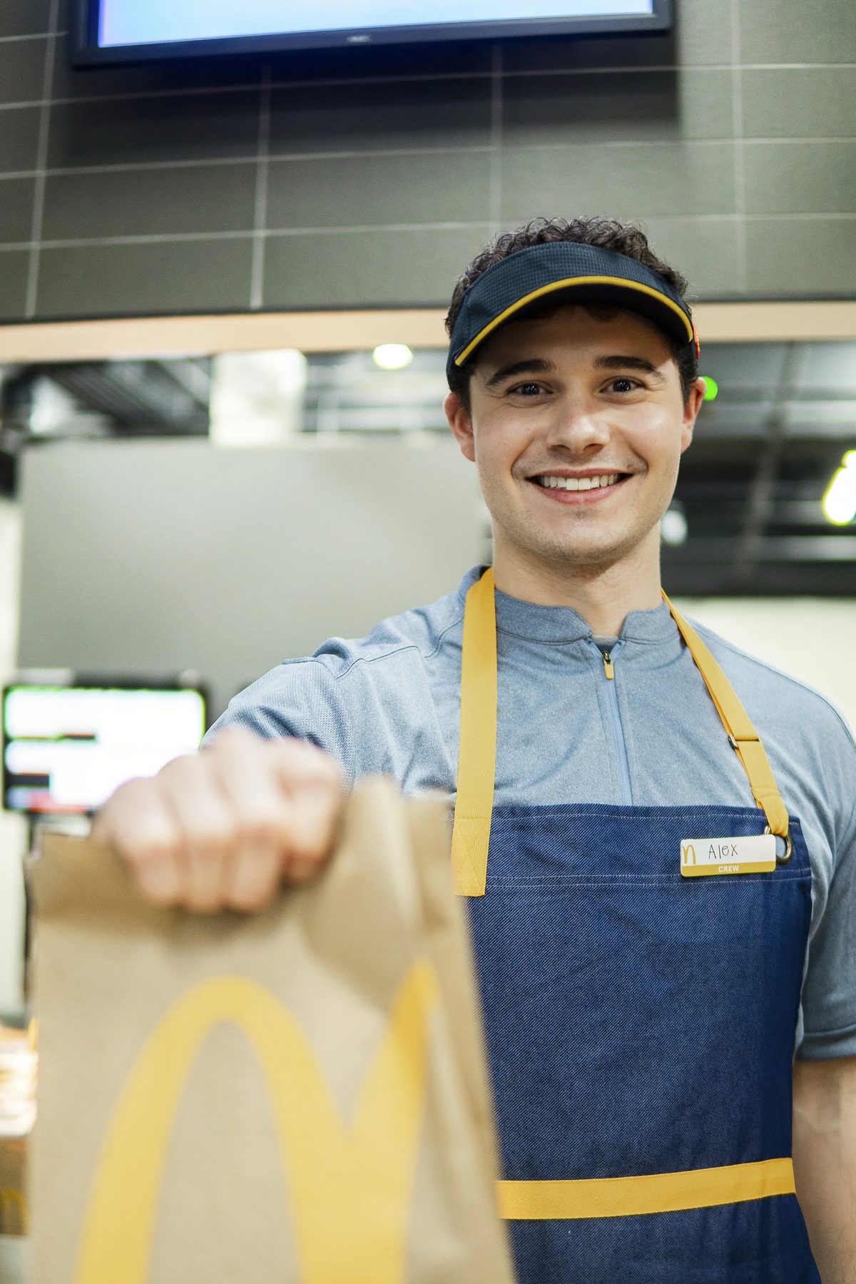 A smiling young man wearing a blue uniform, yellow apron, and a name tag that reads 'Alex' inside a McDonald’s restaurant.