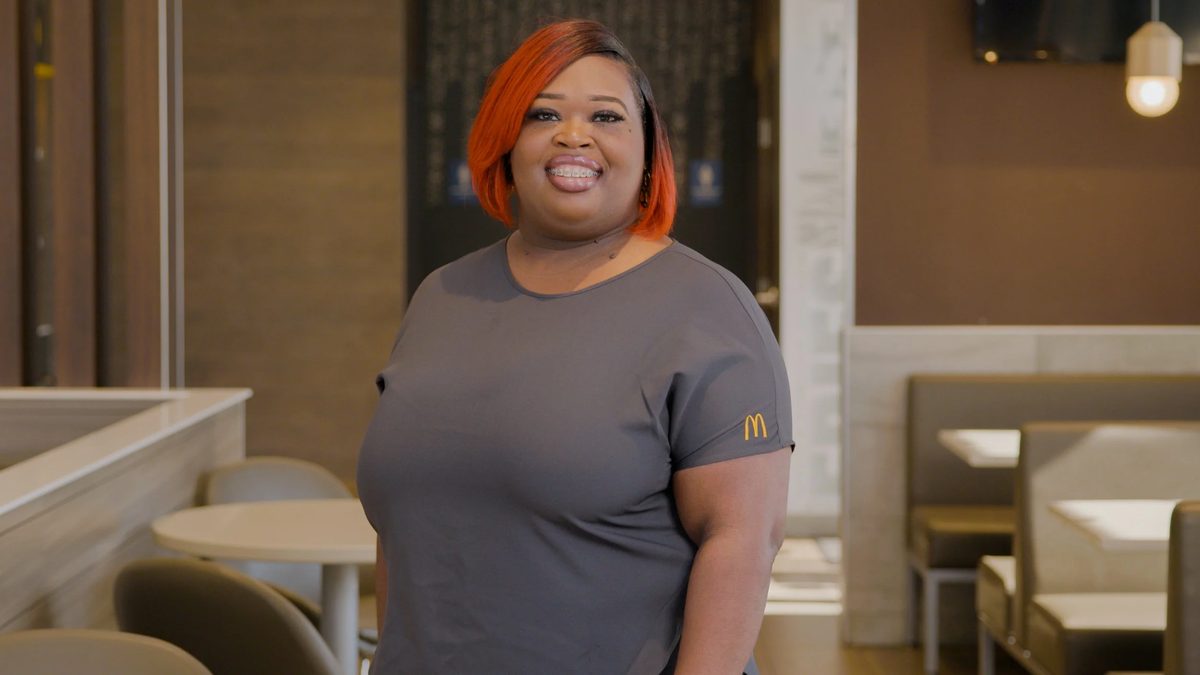 A woman with orange hair wearing a gray McDonald’s uniform standing inside a restaurant.