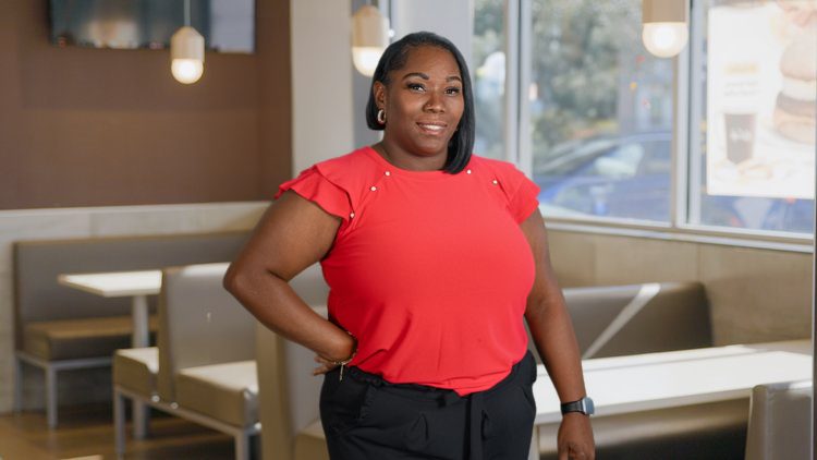 A woman in a red blouse and black pants standing near a window inside a McDonald’s restaurant, smiling at the camera.