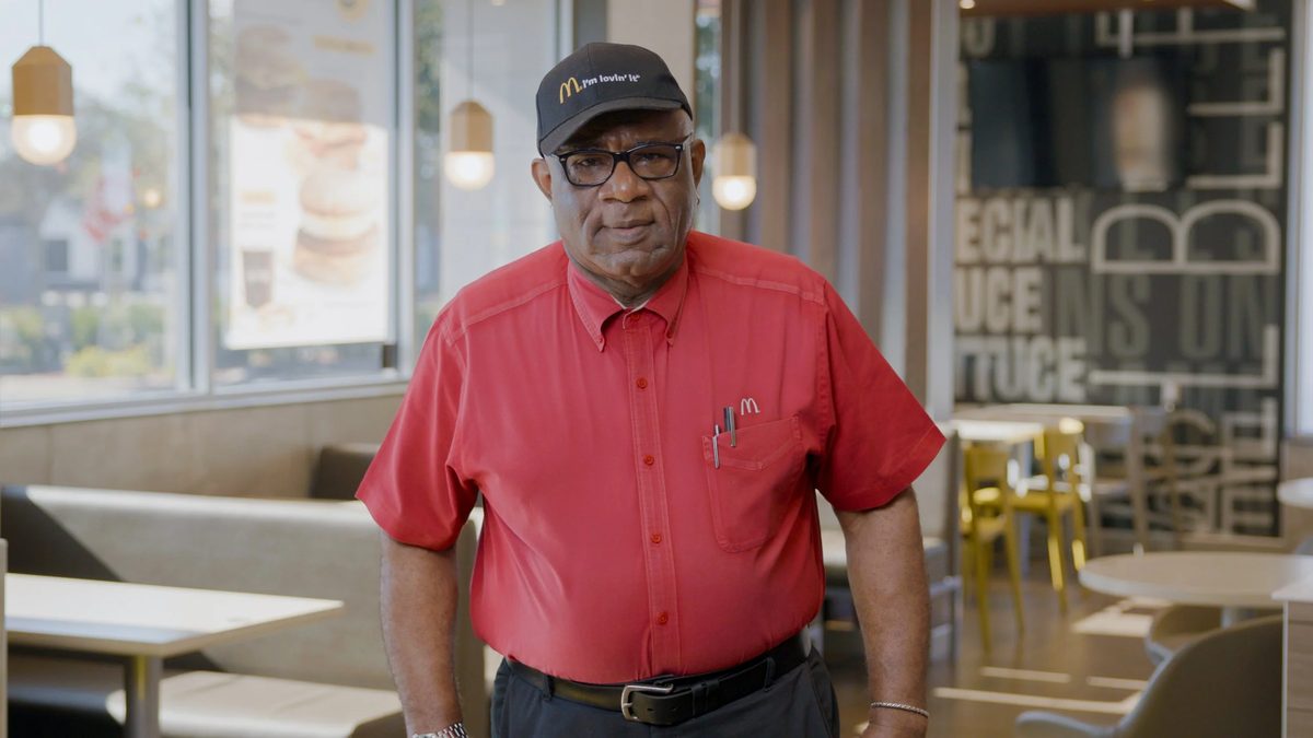 A man in a red McDonald’s uniform shirt and black cap standing inside a restaurant.