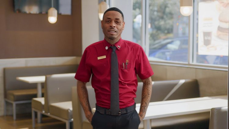 A man in a McDonald’s uniform standing inside a restaurant, smiling with his hands in his pockets.