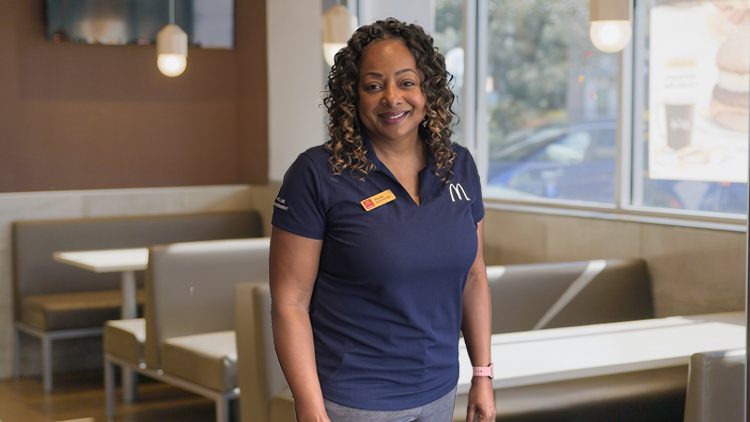A woman in a navy McDonald’s uniform standing inside a restaurant with booths and a large window.