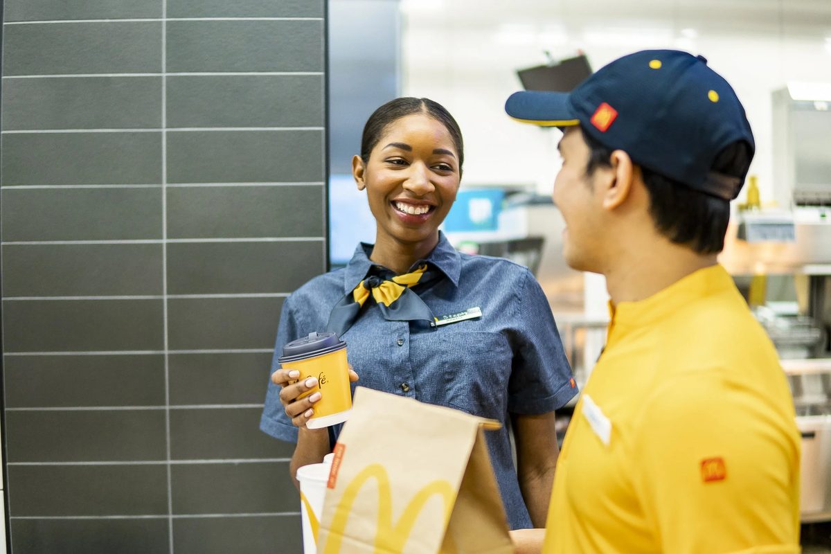 A smiling woman in uniform with a name badge talking to a guest in a yellow uniform inside a McDonald’s restaurant.