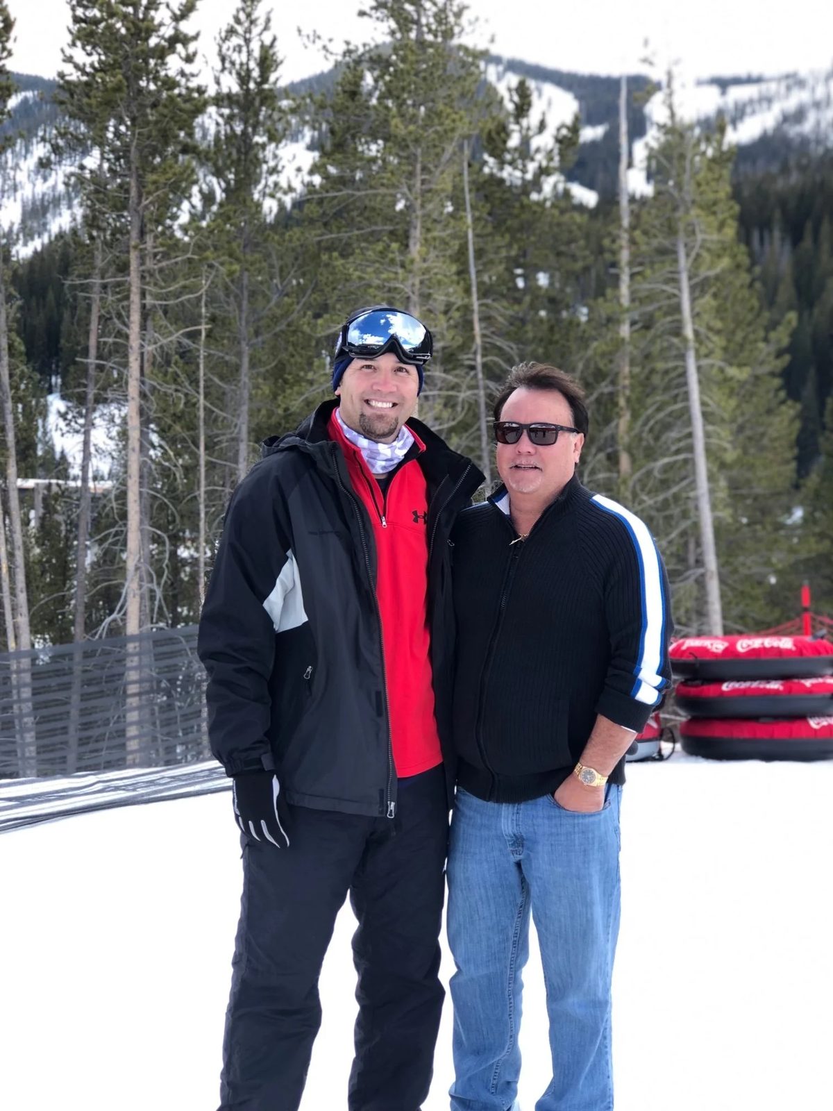 Two men standing outdoors in a snowy mountain landscape with trees, wearing winter clothing.