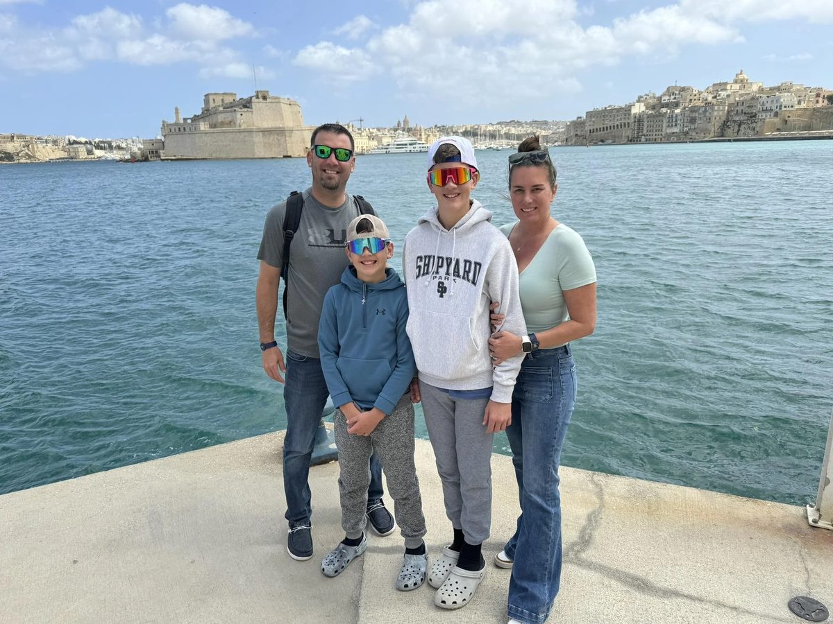 A family of four standing on a concrete pier near the water with boats and historic buildings in the background.