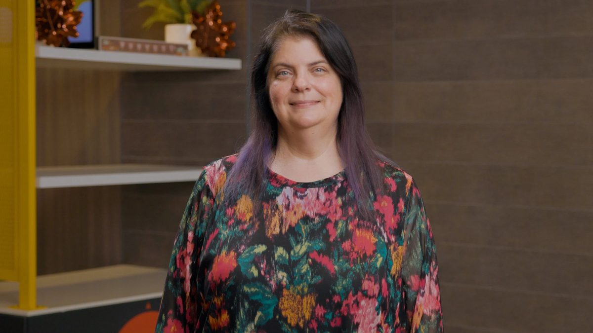 A woman with dark hair and purple streaks wearing a colorful floral top, standing indoors with wooden panel walls.
