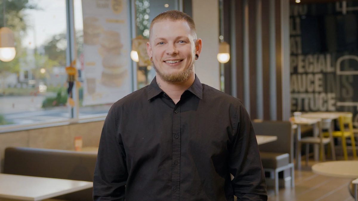 A smiling man with short hair and a beard, wearing a black shirt, standing inside a restaurant.