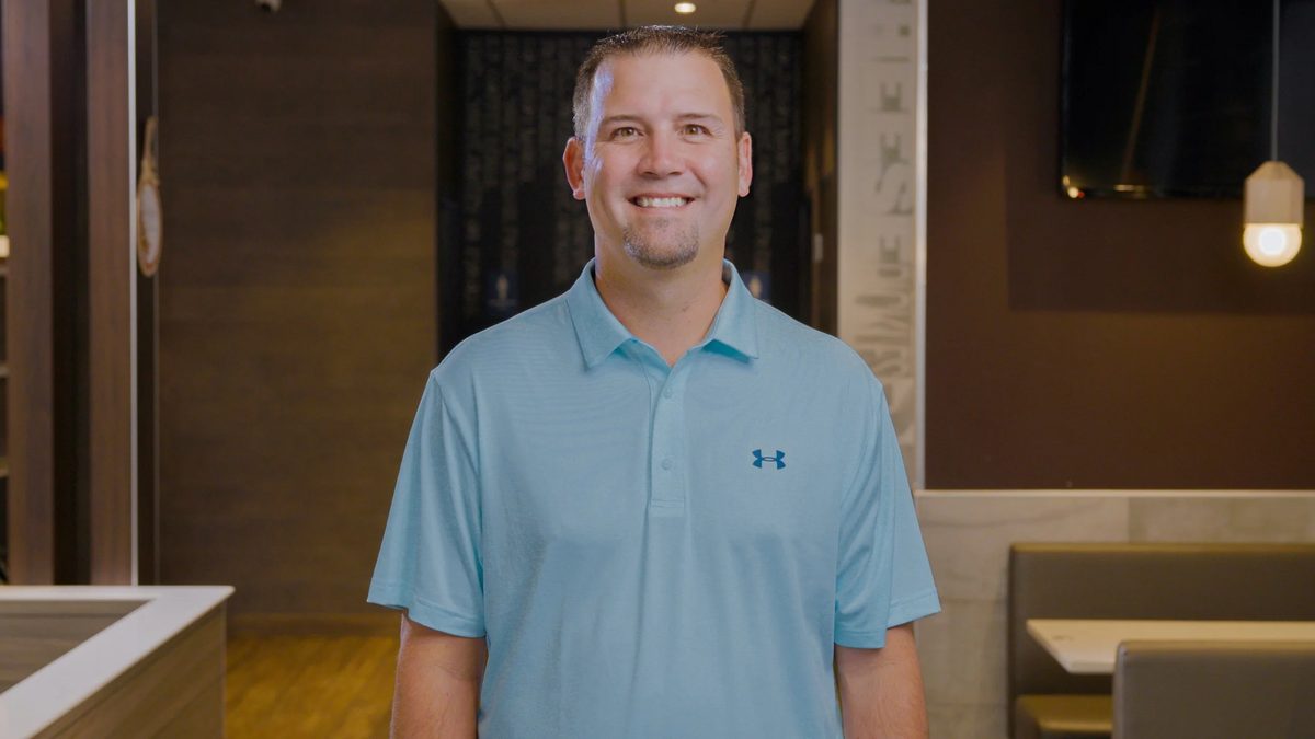 A smiling man wearing a light blue polo shirt standing inside a restaurant with wooden decor and hanging lights.
