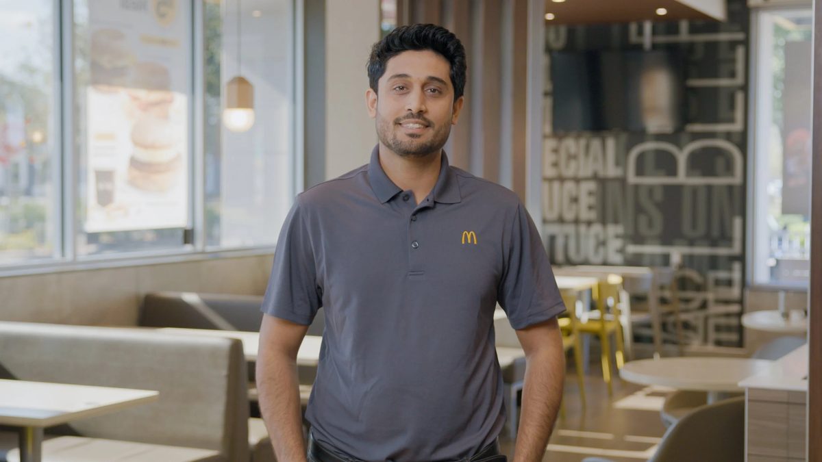 A McDonald’s employee in a gray uniform with a yellow arches logo, smiling inside a restaurant.