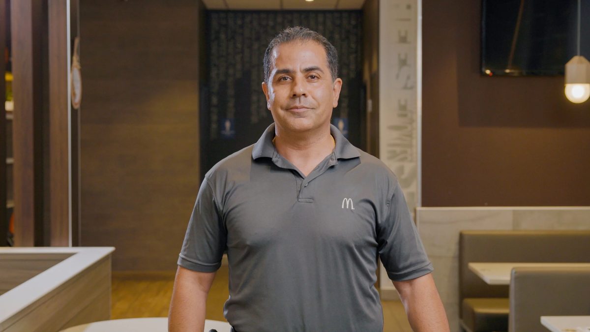 A man in a dark gray McDonald’s uniform standing inside a restaurant with wooden booths and a chalkboard menu behind him.
