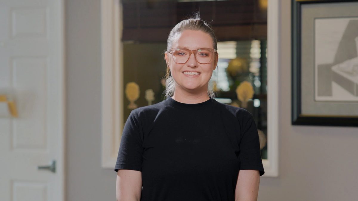 A woman with glasses and a black shirt standing indoors, smiling at the camera.