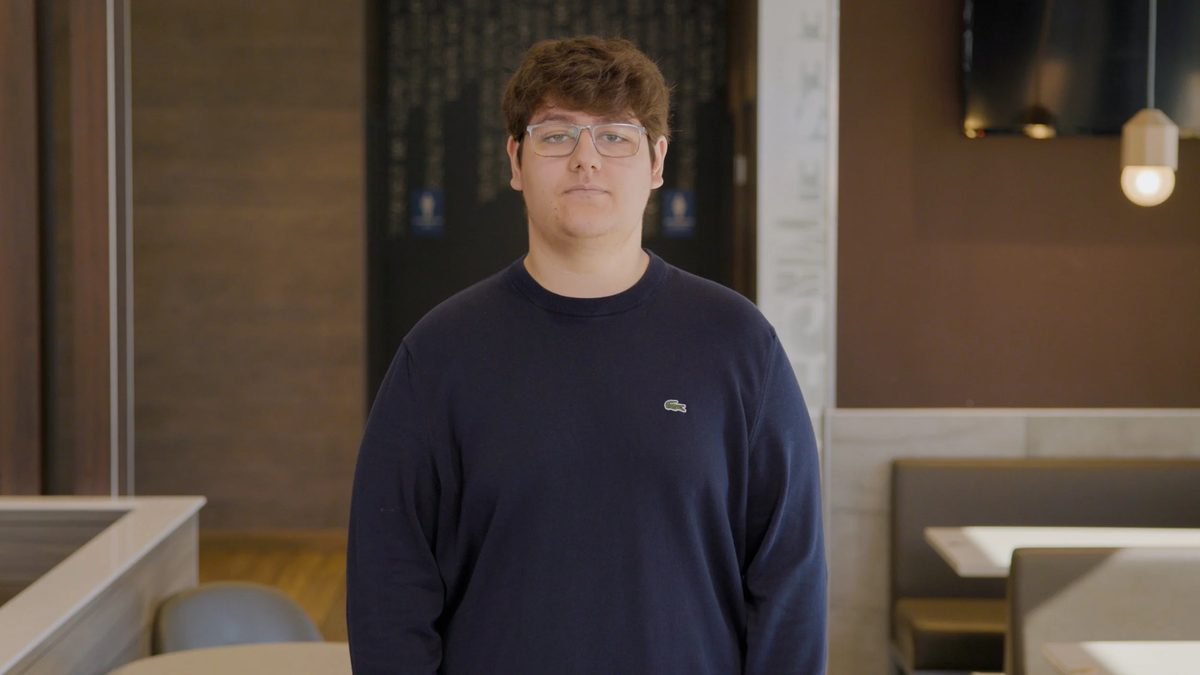 A young man with glasses and brown hair wearing a navy sweatshirt, standing in a modern cafe with brown walls.