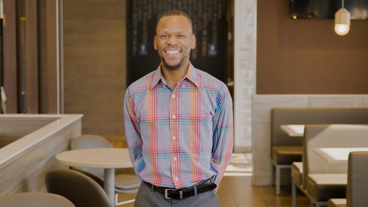 A smiling man with a short beard wearing a colorful plaid shirt and black belt, standing in a McDonald’s dining room with booths and tables behind him.